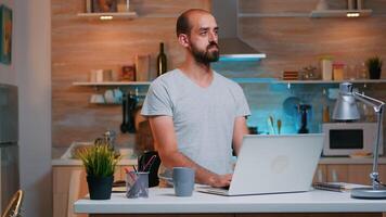 Man thinking at next project while working from home reading the tasks looking on laptop sitting in home kitchen. Busy employee using modern technology network wireless doing overtime reading writing, searching photo
