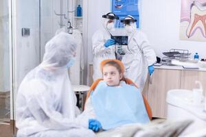 Dentist team analyzing kid radiography dressed in protection gear against coronavirus. Stomatologist during covid19 wearing ppe suit doing teeth procedure of child sitting on chair. photo