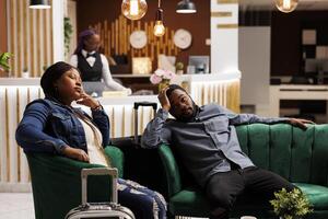 Tired African American man and woman having jet lag symptoms, resting with luggage in hotel lobby. Exhausted tourists feeling unwell after long-distance travel, waiting for check-in procedure photo