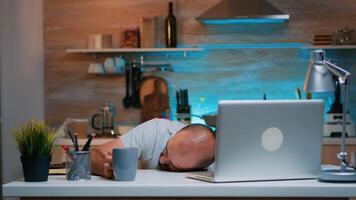 Overworked man sleeping on kitchen desk working remotely late hours at home. Busy exhausted remote employee napping on chair waking up working on laptop using modern technology network overtime photo