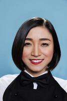 Smiling young attractive asian woman wearing waitress black and white uniform with bow tie closeup portrait. Cheerful woman receptionist looking at camera with positive facial expression photo