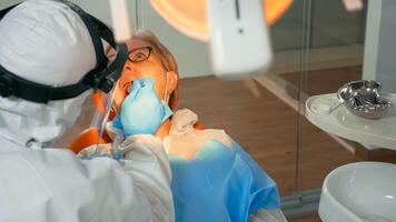 Woman on dental chair in dentist surgery treated with dental drill during covid-19 pandemic. Orthodontist lighting the lamp wearing face shield, protection suit, mask and gloves talking with patient photo