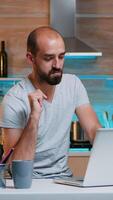 Remote man entrepreneur having video conference with colleagues using laptop sitting in the kitchen late at night. Busy employee using modern technology network wireless reading writing, searching photo