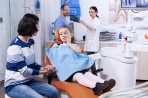 Little girl in dentist office showing parent where tooth hurts touching face with painfull expression. Child with her mother during teeth check up with stomatolog sitting on chair. photo