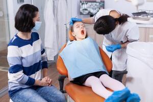 Little kid with mouth open wearing bib sittingon dental chair and dentist looking for caries Dentistry specialist during child cavity consultation in stomatology office using modern technology. photo