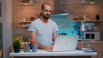 Man discussing on video call working from home sitting in kitchen looking on laptop. Busy focused employee using modern technology network wireless doing overtime for job reading writing, searching photo