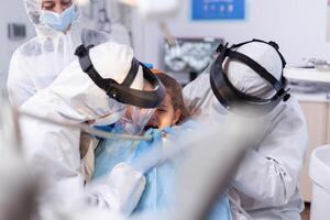 Dentist making tooth caries treatment using drill and dental mirror to little girl Stomatology team wearing ppe suit during covid19 doing procedure on child teeth. photo