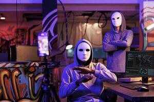 Anonymous hackers recording ransom scam on smartphone while doing illegal activities. Criminals streaming threat online and committing cyberattack in abandoned warehouse at night photo