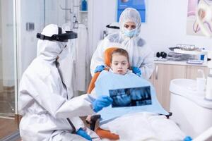 Little girl and mother wearing ppe suit looking at dental radiography in dentist office during covid19 pandemic. Stomatolog in protectie suit for coroanvirus as safety precaution holding child teeth x-ray during consultation. photo