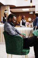 Focused black guy freelancer sitting in hotel lobby with laptop working remotely while traveling. Young African American man traveler using computer during business trip, waiting for check-in photo