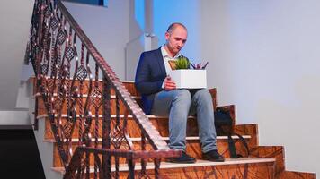 Unemployed businessman sitting on office stairs in depression with box of stuff, lost his business. Group of professional successful businesspeople working in modern financial building. photo