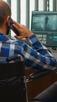 Disabled freelancer videographer talking on phone during editing a video project creating content sitting in wheelchair in modern company office. Creator blogger working from photo studio.