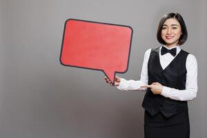 Smiling asian waitress in uniform holding red empty chat bubble and pointing with finger portrait. Cheerful woman receptionist showing blank message banner and looking at camera photo