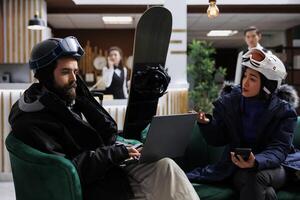 In hotel lobby young guests on couch plan skiing and snowboarding activities with digital devices. Couple using technology for online check-in at ski resort. Snowboard helmets and laptop available. photo