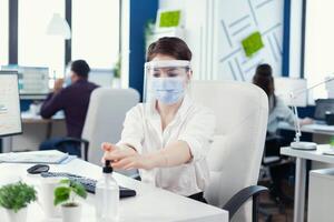 Entrepreneur applying hand sanitizerd uring global pandemic with coronavirus wearing face mask and face shield. Businesswoman in new normal workplace disinfecting while colleagues working in background. photo