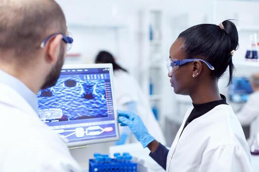 Serious african scientist standing using computer for medical experiments with help from colleague. Multiethnic team of medical researchers working together in sterile lab wearing protection glasses and gloves. photo