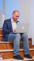 Overworked tired businessman at workplace working on deadline for job project using notebook sitting on stairwell. Confident corporate entrepreneur using laptop doing overtime in business building. photo