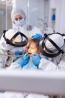Little girl at dentist's appointment having a painfull tooth treatment dressed in protective suit against covronavirus. Stomatology team wearing ppe suit during covid19 doing procedure on child teeth. photo