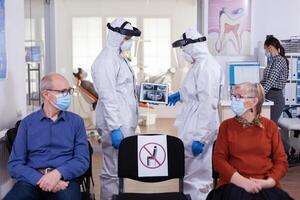 Man discussing with nurse in dental reception wearing protection suit against coronavirus, elderly patients waiting in reception keeping distance. Concept of new normal dentist visit in outbreak. photo