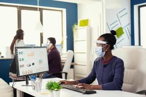 African woman at workplace working on computer looking at graph wearing face mask against covid19. Multiethnic team in new normal business financial office checking reports, analysing datas looking at desktop. photo
