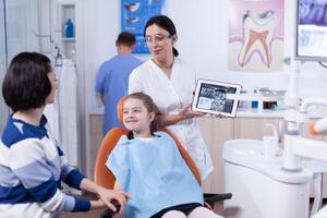 Excited little girl in dentist cabinet for periodic check up looking at parent. Stomatologist explaining teeth diagnosis to mother of child in health clinic holding x-ray. photo