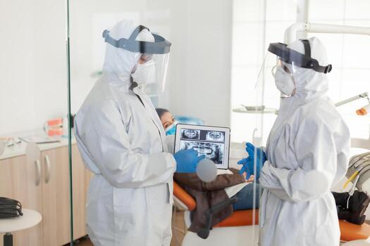 Dentist doctors in overall using tablet explaining dental x ray in stomatologic office during coronavirus. Man wearing face shield and mask showing to nusrse radiography using digital device photo