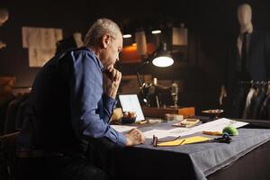 Elderly suitmaker overthinking comissioned business suit required by customer. Old experienced couturier working on sartorial bespoken clothing designs in tailoring studio workspace photo