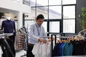Asian employee arranging casual wear items in shopping centre, preparing store for opening. Stylish worker checking clothes fabric and price, putting merchandise on hangers in modern boutique photo