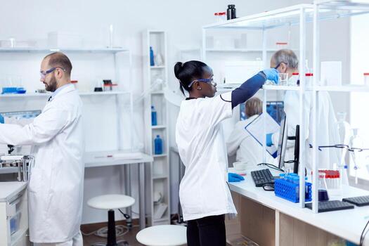 African chemist in professional laboratory taking a glass flask with solution from shelf. Multiethnic team of researchers working in microbiology lab testing solution for medical purpose. photo