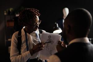 Experienced fashion designer showing couturier sketch drawing of wedding dress she wants him to customize. African american dressmakers working on stylish bespoke sartorial attire photo