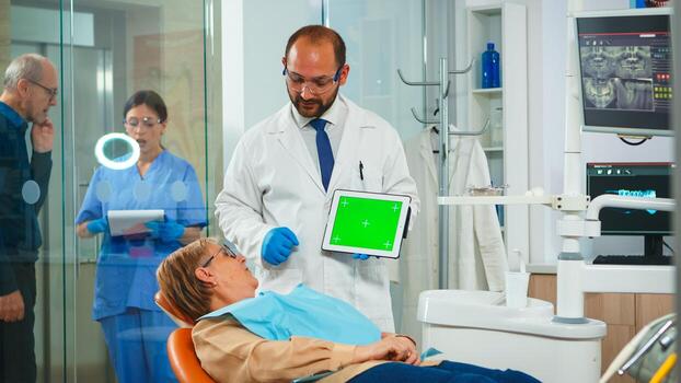 Dentist pointing at green screen display while patient lying on dental chair. Man explaining using monitor with green chroma key isolated chroma pc key mockup greenscreen display mock-up touchscreen photo