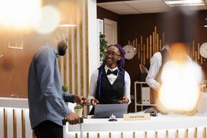 Smiling friendly African American woman receptionist talking with man guest at hotel reception. Black guy tourist standing with baggage at front desk doing check-in, receiving tourist information photo