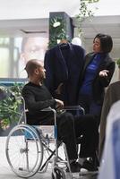 Clothing store assistant and arab man customer in wheelchair examining suit on hanger together and chatting. Mall seller and buyer with disability exploring apparel options photo