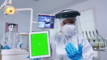 Doctor in ppe suit showing tablet with green screen, explaining dental radiography and diagnosis for teeth infection. Stomatology specialist in coverall pointing at mockup, copy space, chroma display photo