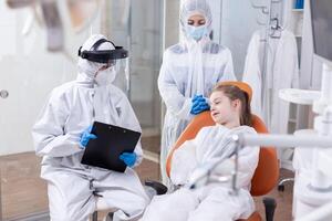 Pediatric dentist talking with little girl about oral hygine in the course of coronavirus pandemic. Stomatologist during covid19 wearing ppe suit doing teeth procedure of child sitting on chair. photo