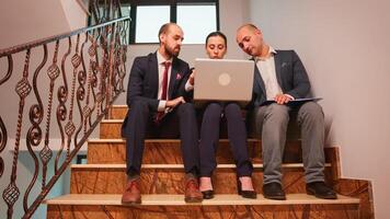 Overworked business colleagues doing overtime sitting on stairs using laptop and tablet, analysing difficult finance project. Company manager and office executive on stairs in corporate building. photo