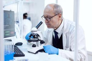 Senior man working in life science laboratory using modern microscope. Chemist researcher in sterile lab doing experiments for medical industry using modern technology. photo
