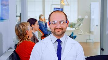 Portrait of stomatologist smiling at camera being in dental office while patients waiting him in background. Dentistry doctor looking on webcam sitting on chair in stomatological clinic. photo