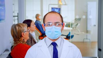 Portrait of tired stomatologist wearing protective mask at camera being in dental office while patients waiting him in background. Dentist looking on webcam sitting on chair in stomatological clinic. photo