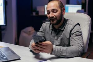 Entrepreneur writing text on smartphone sitting at desk in empy workplace. Businessman using his cell to text message while working late at night in the office to finish a deadline. photo