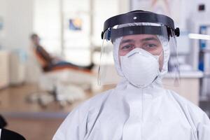 Portrait of tired doctor in dental office looking on camera wearing overall and face shield sitting on chair in waiting room clinic. Concept of new normal dentist visit in coronavirus outbreak. photo