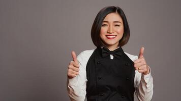 Smiling waitress giving thumbs up sign in front of camera, working in a five star restaurant to serve tables. Asian server with apron showing approval with okay symbol in studio, agreement. Camera B. photo