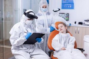 Dental specialist holding clipboard during coronavirus outbreak and little girl sitting on chair. Stomatologist during covid19 wearing ppe suit doing teeth procedure of child sitting on chair. photo