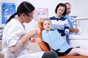 Child learning how to use tootbursh from young dentist in Little girl and mother listening stomatolog talking about tooth hygine in dentistiry clinic holding jaw model. photo