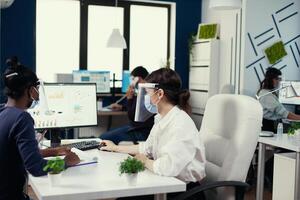 Diverse coworkers looking at computer screen with economic graph wearing face mask against covid19. Multiethnic team working in company with new normal respecting social distance during global pandemic with coronavirus. photo