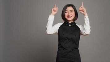 Asian woman pointing at something upwards in studio, indicating direction above her head. Waitress server with apron showing up indication over grey background, table service. Camera A. photo