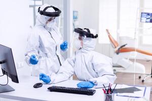 Dentist doctor in ppe suit discussing with nurse in stomatology clinic. Medicine team wearing protection gear against coronavirus pandemic in dental reception as safety precaution. photo