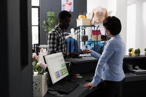 Stylish customer paying for fashionable clothes with credit card using payment terminal during fashion shopping session in modern boutique. African american man buying formal wear photo