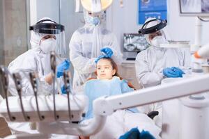 Mother dressed in ppe suit comforting daughter in the course of dental treatment. Stomatology team wearing ppe suit during covid19 doing procedure on child teeth. photo