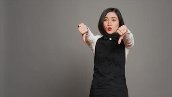 Asian restaurant hostess showing thumbs down and dislike sign, expressing negativity and disapproval in studio. Displeased waitress working at a diner and serving customers. Camera A. photo